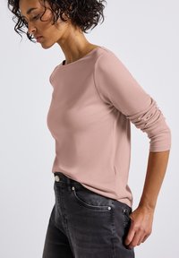 Langärmliges, runde-Halsausschnitt-Oberteil aus weichem pinken Stoff, kombiniert mit dunkelgrauen Jeans. Das Oberteil hat einen figurschmeichelnden Schnitt und leicht aufgerollte Ärmel.