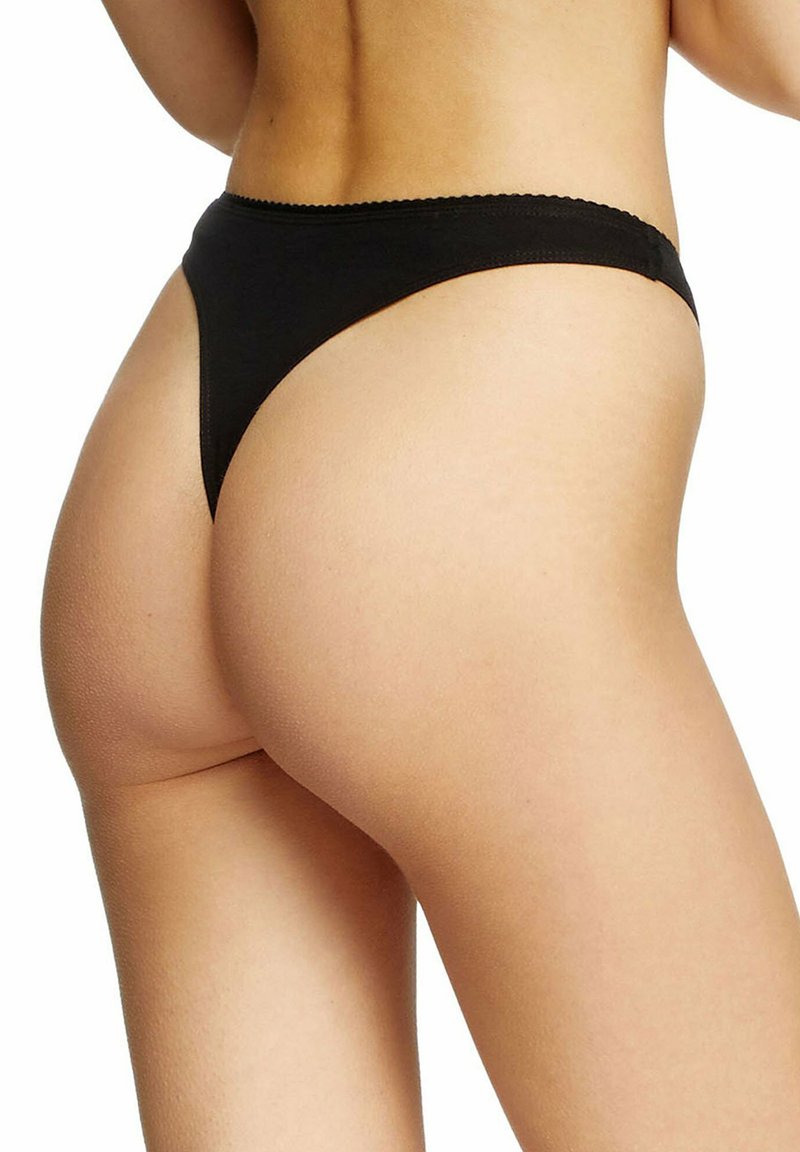 Mujer con ropa interior estilo tanga negra, espalda y parte superior de los muslos visibles contra un fondo blanco.