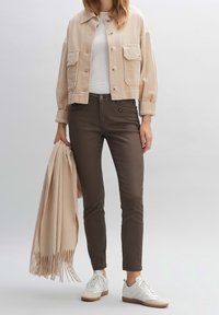 Vaikka beige corduroy-takki, valkoinen t-paita, ruskeat kapeat housut ja valkoiset tennarit. Pitää käsissään vaaleanruskeaa fringe-siteitä.