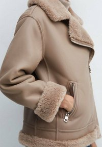 Beżowa skórzana kurtka z puszystym obszyciem ze skóry owczej na kołnierzu, mankietach i dolnym brzegu, z zasuwaną kieszenią, do której ręka jest wsunięta.