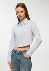 Camicia azzurra chiara a maniche lunghe, con taglio corto, realizzata in tessuto morbido, dotata di colletto classico e chiusura con bottoni.