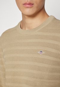 Beiger gestreifter Strickpullover mit geripptem Rundhalsausschnitt; verfügt über ein kleines gesticktes Logo auf der Brust. Weiche Textur und lässige Passform.
