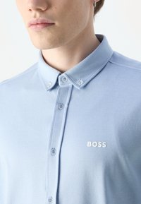 Jasnoniebieska polo z teksturowanej tkaniny z kołnierzykiem zapinanym na guziki i białym logo "BOSS" na klatce piersiowej. Zawiera małe białe guziki.