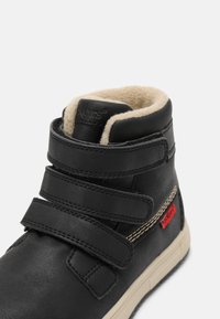 Stivale in pelle nera con interno in pile beige, tre cinturini regolabili in velcro e dettaglio del logo rosso. Presenta una punta tonda e una suola testurizzata.