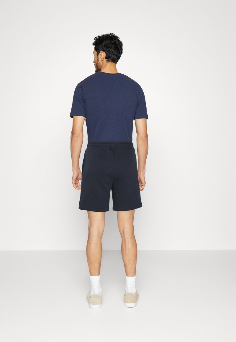 Navyblaue Baumwoll-T-Shirt kombiniert mit schwarzen Shorts, die einen elastischen Bund haben. Weiße Socken und hellbraune Schuhe vervollständigen das Outfit.
