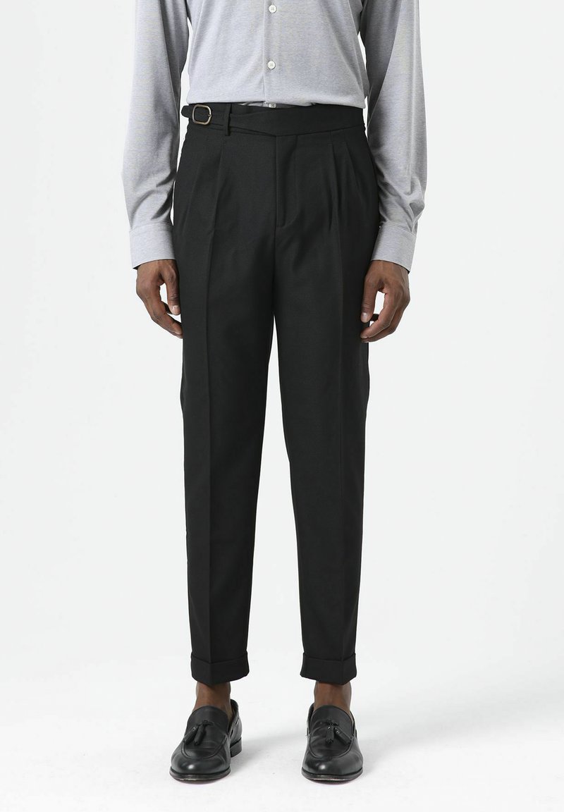 Zwarte, op maat gemaakte broek met plooien, gespsluiting en opgerolde manchetten. Gecombineerd met een lichtgrijs shirt met lange mouwen en zwarte loafers.