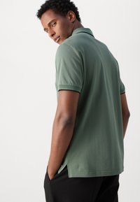 Groene polo met korte mouwen, contrasterende kraag en zijslitsen, gemaakt van zacht materiaal. Model met handen in de zakken, gezien van achteren.