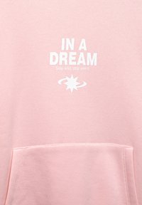 Ružová mikina s bielym potlačeným textom "IN A DREAM" a "Stay wild, stay weird," so grafickým prvkom hviezdy a dizajnom prednej vrecka.