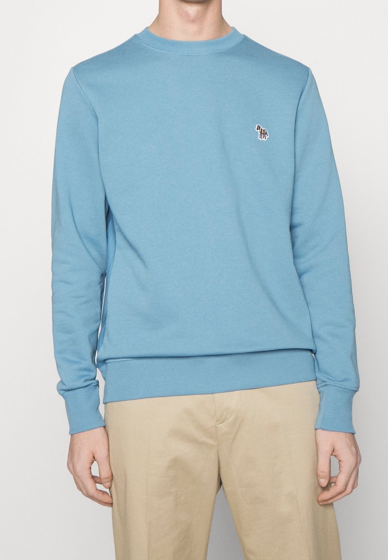 Homme portant un sweat-shirt à manches longues bleu clair avec un petit logo zèbre multicolore sur la poitrine et un pantalon beige, debout devant un fond uni.