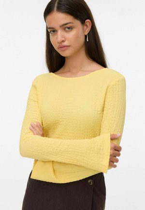 Vrouw met lang, donker haar, gekleed in een textuur geel lange mouwen top en een donkere rok, met gekruiste armen en hangende pareloorbellen, tegen een witte achtergrond.
