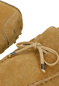 Tan-farbene Wildleder-Loafer verfügen über eine abgerundete Spitze, dünne Lederbänder mit metallischen Enden und ein strukturiertes Obermaterial mit Nähdetails.