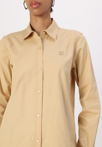 Beige pika-särgid, millel on klassikaline krae, esi-nupud ja tikitud "HUGO" logo rinnal. Sile tekstuur.