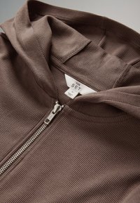 Felpa con cappuccio marrone a coste con cerniera e tessuto morbido. Zip in metallo. Etichetta con la scritta "THE SET," taglia media. Nessun motivo o accento visibile.