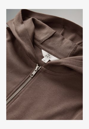 Rudas, dėžuotas megztinis su užtrauktuku, minkšto audinio ir metaliniu užtrauktuku. Etiketėje užrašyta „THE SET“, dydis vidutinis. Matomų raštų ar akcentų nėra.