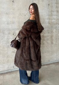 Brun pälsimitation kappa med ett tjockt, strukturerat utseende, kombinerad med en randig off-shoulder topp och utsvängda jeans. Brun handväska som accent.