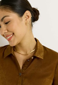 Collar y pendientes de color dorado con diversas formas geométricas. La modelo lleva una camisa de satén marrón con cuello y botones decorativos.