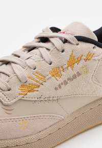 Beige mockask sneaks med gula och vita broderade mönster, diskret snörning, strukturerad gummisula och förstärkt tåhätta.