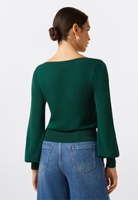 Grüner Strickpullover mit weitem Ausschnitt, langen puffigen Ärmeln und taillierter Passform. Kombiniert mit hochtaillierten blauen Jeans.