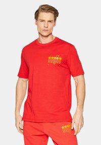 T-shirt di cotone rosso con maniche corte, caratterizzata da un logo grafico giallo e testo nella zona del petto sinistro. Scollo rotondo, vestibilità comoda.