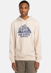 Cream hooded sweatshirt featuring a blue graphic design with text "THE DOERS" and forest elements. Front kangaroo pocket and ribbed cuffs.