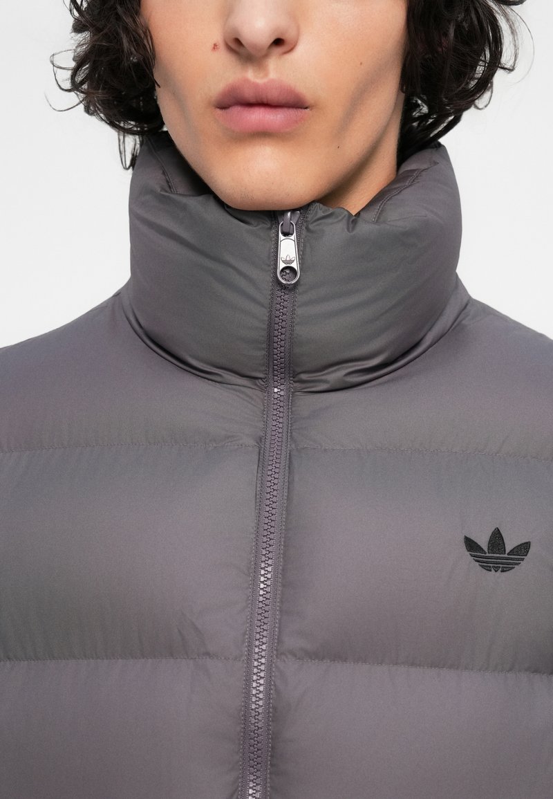 Joven con cabello rizado que lleva una chaqueta acolchada gris de cuello alto con un logo negro de Adidas en el pecho, cremallera completamente cerrada.