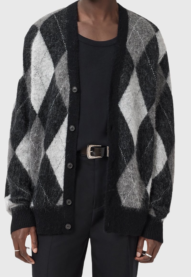 Personne portant un cardigan à motif losange noir et gris sur une chemise noire, associé à un pantalon noir et une ceinture noire avec une boucle argentée.