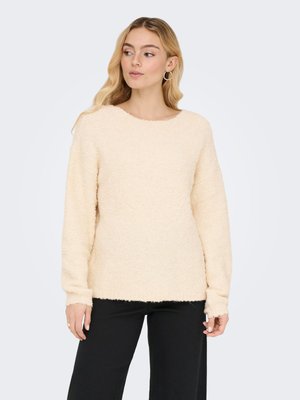 Vrouw met lang blond haar, die hoepel oorbellen, een crèmekleurige textuursweater en zwarte broek draagt, kijkt iets naar links.