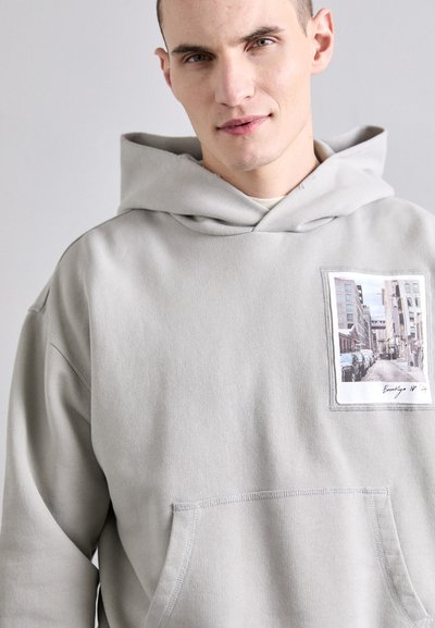 Jasnoszara bluza z kapturem wykonana z tkaniny bawełnianej. Posiada przednią kieszeń oraz naszywkę ze zdjęciem miejskiej sceny po lewej stronie.