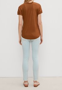 Braunes Kurzarmhemd kombiniert mit hellblauen Skinny Jeans mit rohen Säumen und beigen Sandalen. Das Hemd hat einen abgerundeten Saum und hochgeschlagene Ärmel.