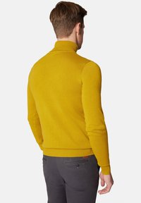 Musztardowy sweter z golfem o slim fit, z prążkowanymi mankietami i dolnym wykończeniem. Nosi się go na ciemnoszarych spodniach, eksponując gładką fakturę dzianiny.