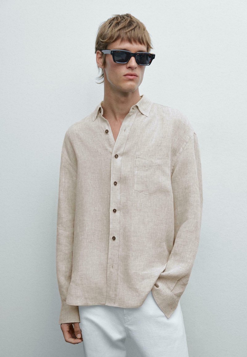 Beige Leinenhemd mit langen Ärmeln, Tasche und braunen Knöpfen. Gestylt mit schwarzen Sonnenbrillen und weißen Hosen.
