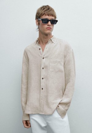 Camicia button-up beige in lino, con maniche lunghe, tasca e bottoni marroni. Abbinata a occhiali da sole neri e pantaloni bianchi.