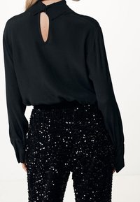 Blusa nera a maniche lunghe con colletto, caratterizzata da un'apertura a foro chiave sul retro. Abbinata a pantaloni fitted neri coperti di paillettes.