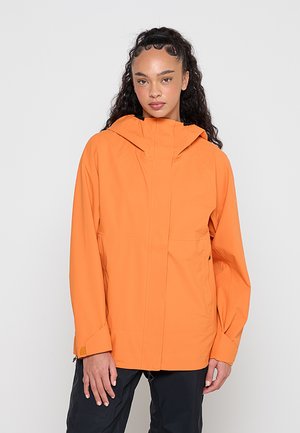 Jeune femme aux longs cheveux bouclés portant une veste à capuche orange et un pantalon noir, debout devant un fond clair uni.