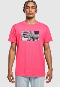 Rožinė medvilninė marškinė, su juodai balta nuotrauka centre, vaizduojančia tatuiruotą ranką, laikantį grandines, o po ja – balti žodžiai „melskis“. 