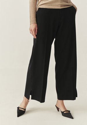 Schwarze, weit geschnittene Hosen mit Seitenschlitzen, kombiniert mit schwarzen spitzen Mules mit Riemen. Weicher, glatter Stoff mit fließendem Design.