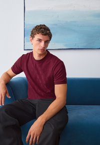 Burgundy textured short-sleeve sweater with geometric patterns, paired with black trousers, seated on a blue couch against a light wall.