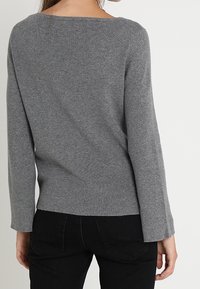 Grå strikket sweater med bred halsudskæring og lange, let klokkeformede ærmer. Blød tekstur, tætsiddende ved taljen, parret med sorte jeans.