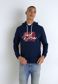 Felpa navy con coulisse bianche, con un design del logo rosso e bianco. Abbinata a jeans chiari strappati di colore blu.
