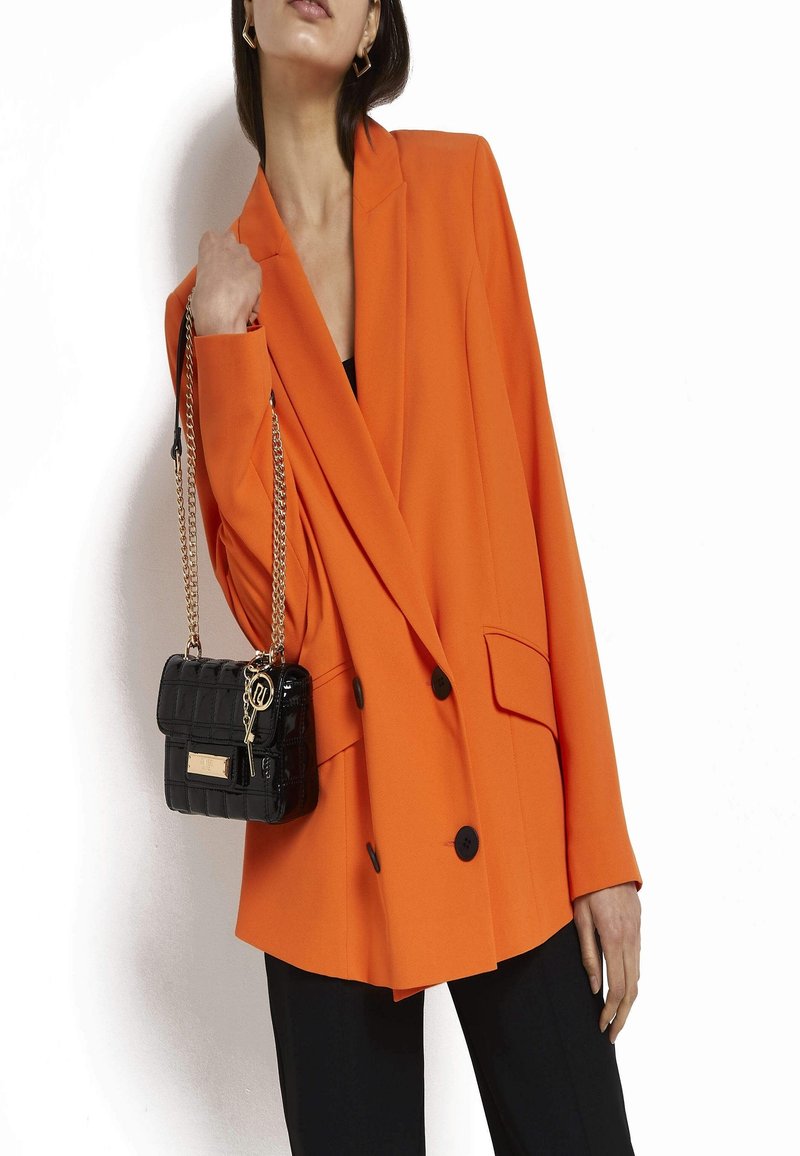 Blazer tailored di un brillante arancione con un design doppiopetto, abbinato a una piccola borsa a tracolla nera trapuntata con una catena dorata.
