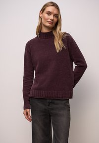 Pflaumenfarbener Strickpullover mit hohem Kragen, geripptem Saum und Bündchen; kombiniert mit dunkelgrauer Hose. Einfacher, strukturierter Stoff ohne Muster.