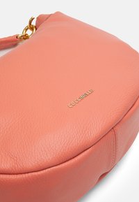 Brzoskwiniowa skórzana torebka o zaokrąglonym kształcie, widoczną fakturą i złotymi akcentami, na których wygrawerowano nazwę marki "Goccinelle".