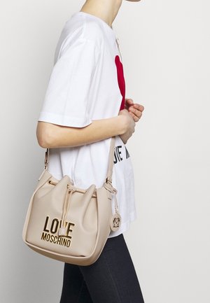 Beige emmer tas met gouden "LOVE MOSCHINO" opschrift, trekkoordsluiting en een decoratief embleem. Draag over een witte shirt met een rood hartontwerp.