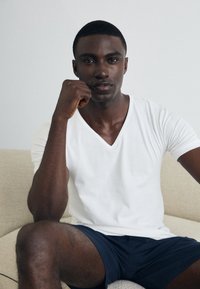 T-shirt blanc à col en V sur une personne assise, associé à un short bleu marine. Texture en coton lisse, coupe décontractée. Teint de peau foncé, traits du visage bien définis.
