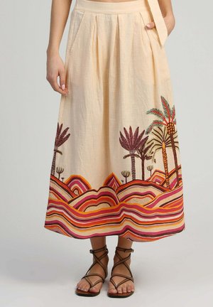 Jupe midi beige avec des plis, ornée de motifs colorés de palmiers et de paysages rayés le long de l'ourlet, portée avec des sandales à brides marron.