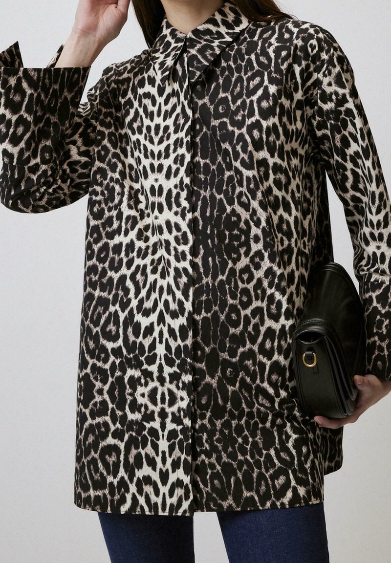 Femme portant une chemise léopard à manches longues boutonnée, tenant un sac à main en cuir noir, visible du cou jusqu'aux cuisses.