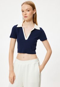 Polo shirt cropped blu navy con colletto bianco a contrasto e maniche corte, caratterizzato da un design a V e texture ribbata. Abbinato a pantaloni bianchi.
