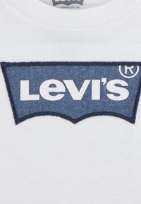 Valge t-särk, millel on suur sinine teksakangast plaastritükk, mis on kujult nagu Levi'si logo, valge tekstiga ja servade ümber detailne õmblus.