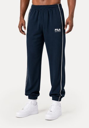 Mand iført marineblå FILA joggingbukser med hvide sidelister, hvide sokker og hvide sneakers, vist fra talje til fødder.