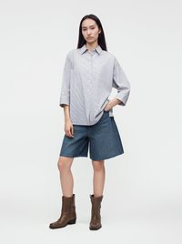 Femme debout, main dans la poche, portant une chemise ample à rayures, un short en jean longueur genou et des bottines marron sur fond uni.